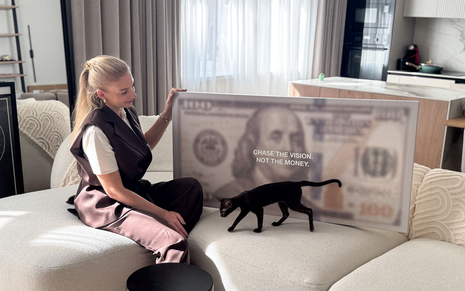 Woman sitting on a couch with a large dollar bill canvas art and a black cat in a modern living room.
