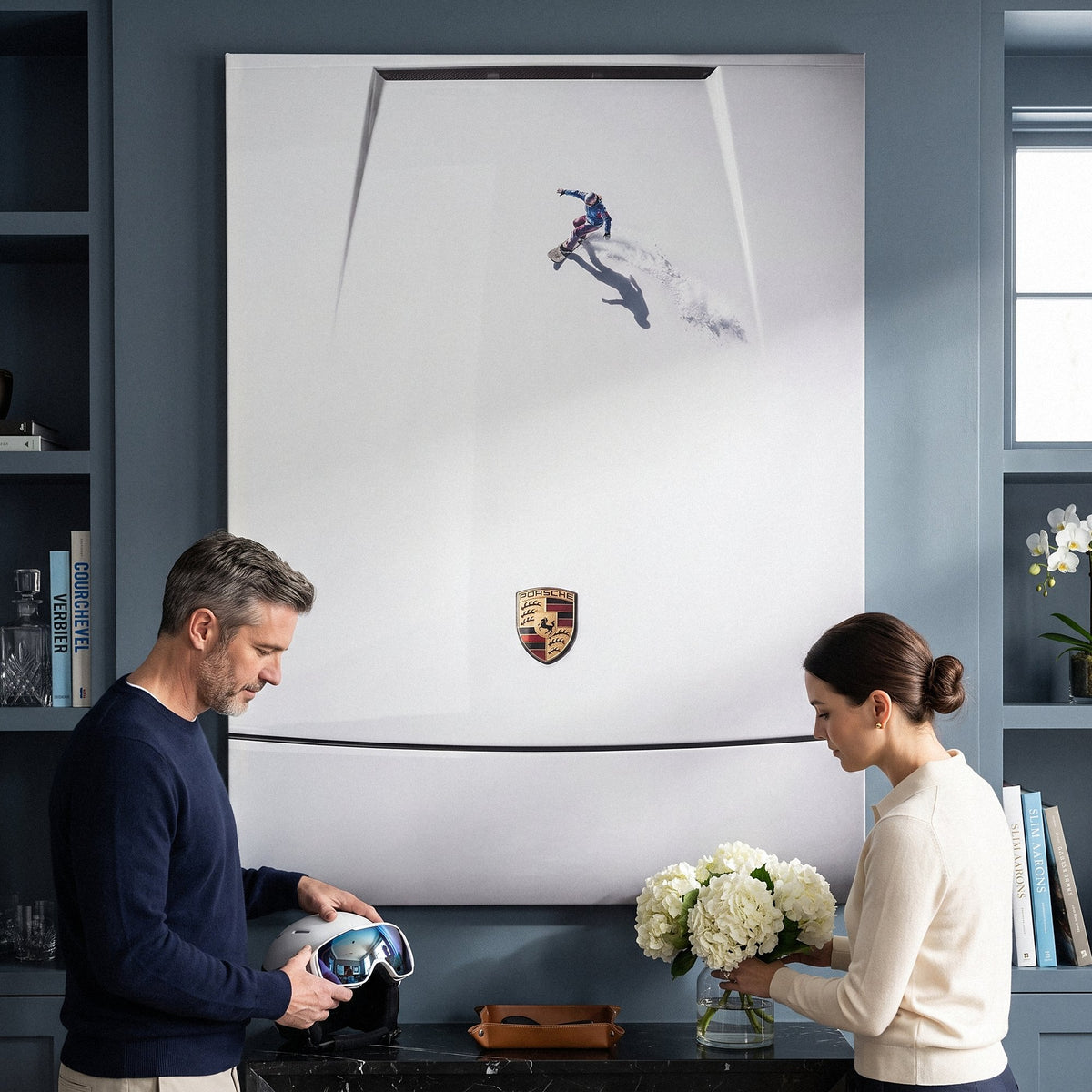 Couple below the Car Boarding canvas in a slate blue room, he holds a ski helmet and she arranges white hydrangeas
