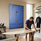 Man waxing a longboard on stands below the Car Surfing canvas, with surfboards and a wetsuit on the wall