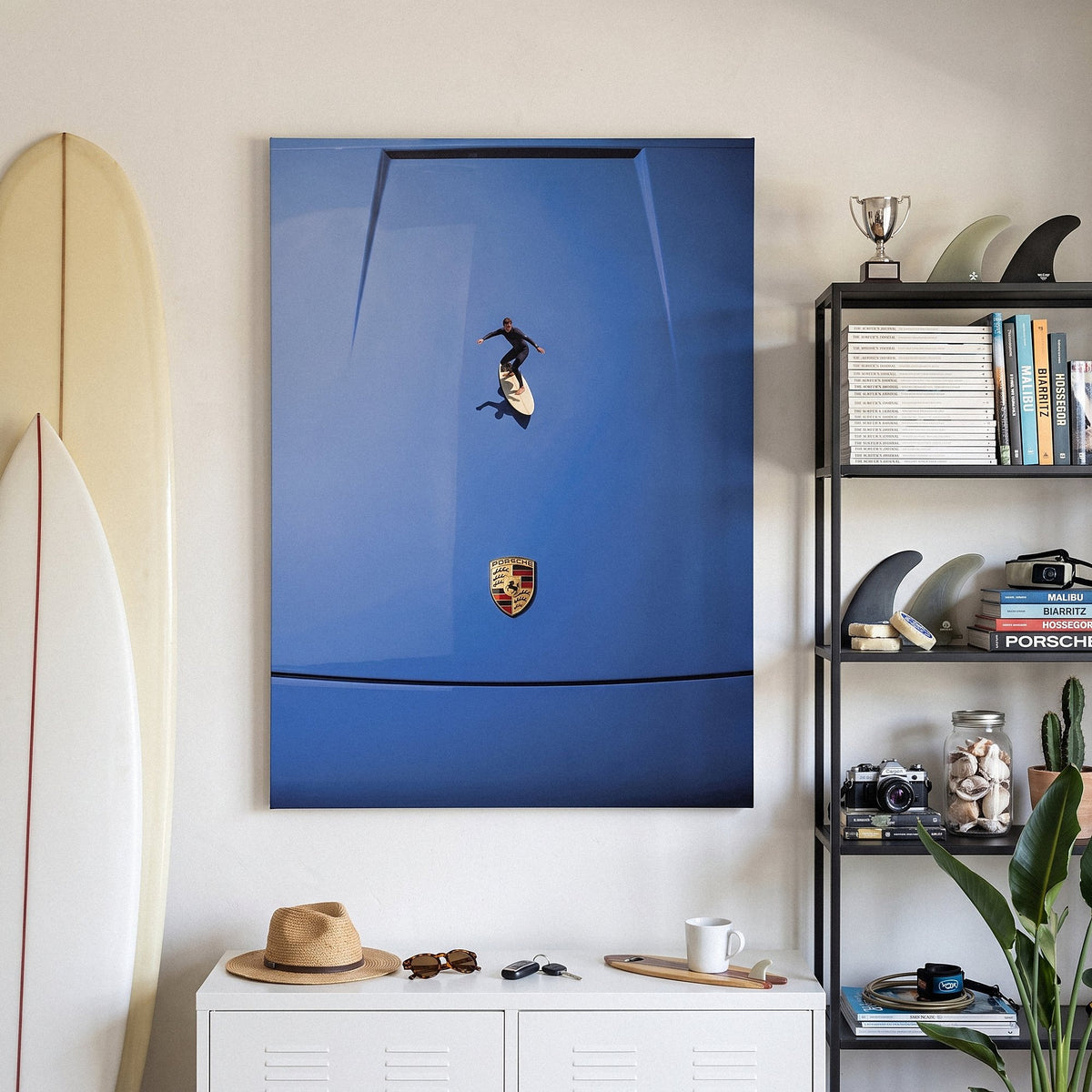 Car Surfing canvas in a surfer's room with longboards, a black shelf of fins and surf magazines, and a straw hat on a white cabinet