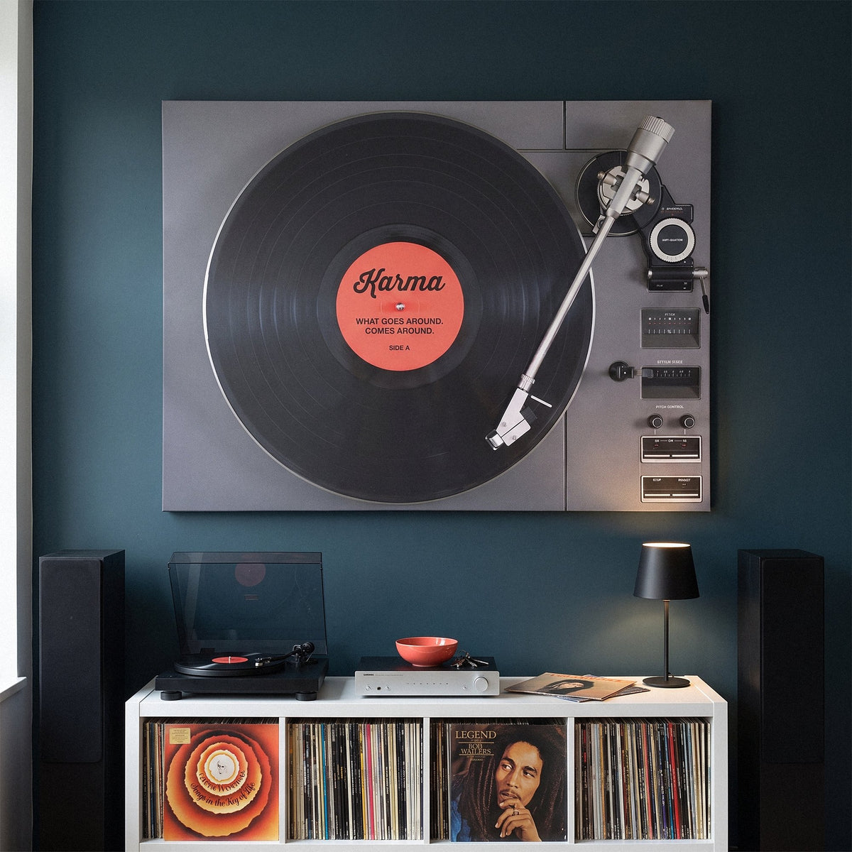 Karma turntable canvas above a white record console between tall floor speakers on a petrol navy wall